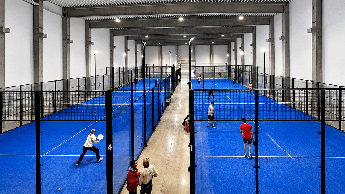 In einer großen Halle spielen mehrere Personen auf vier blauen Padelcourts unter hoher Betondecke.