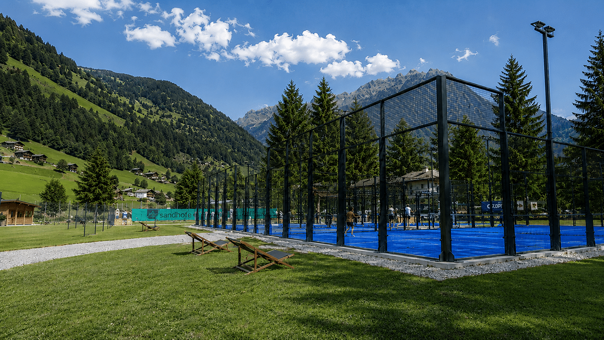 Ein blaues Padel-Feld mit hohen Glaswänden liegt in einer grünen Alpenlandschaft zwischen Tannen und Bergen.